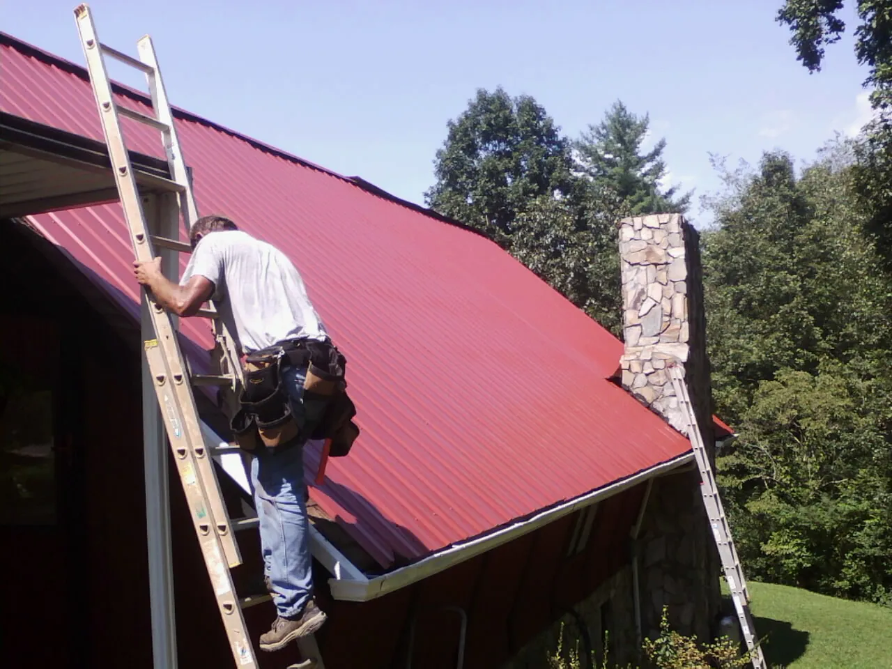 Roofer on a ladder beside a red metal roof and stone chimney — Soffit & Fascia Repair by Roofing Brothers in Maynard