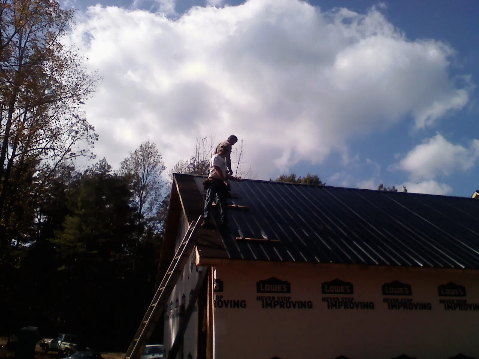 Roofer fastening dark metal panels high against a cloudy sky, from a Roof Inspection job in Maynard
